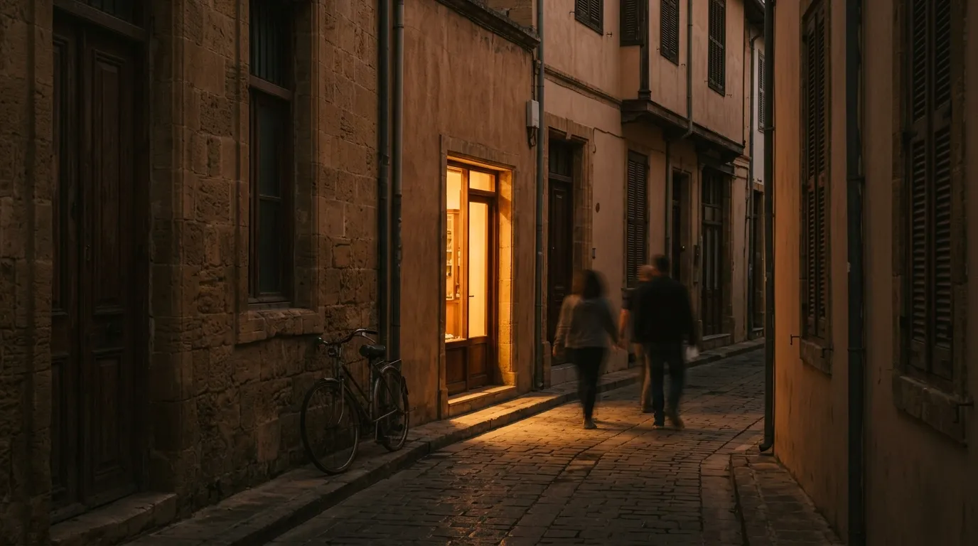 A warmly lit shopfront glowing onto a narrow cobbled Nicosia street at dusk, passers-by drawn towards the light